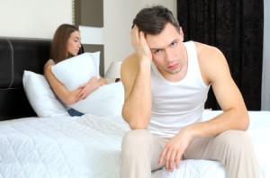 Man sits on the side of the bed, while woman sits in bed holding pillow, both unhappy