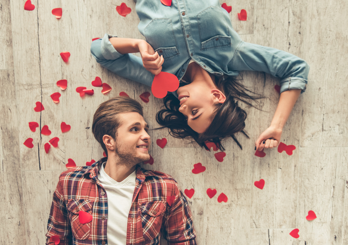 Top view of happy young couple looking at each other and smiling while lying on wooden floor. Girl is holding a red paper heart