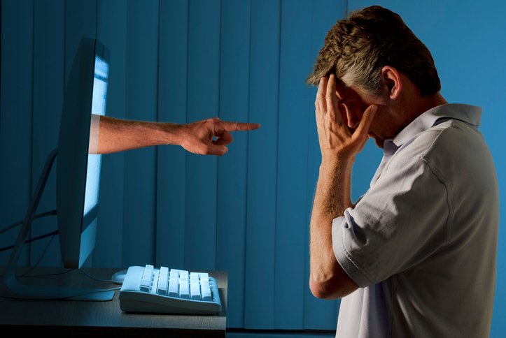 Severely distraught young man sitting in front of a computer with a judgmental hand pointing at him from within the computer monitor which shows the man being either computer bullying bullied or Facebook social media stalking stalked.