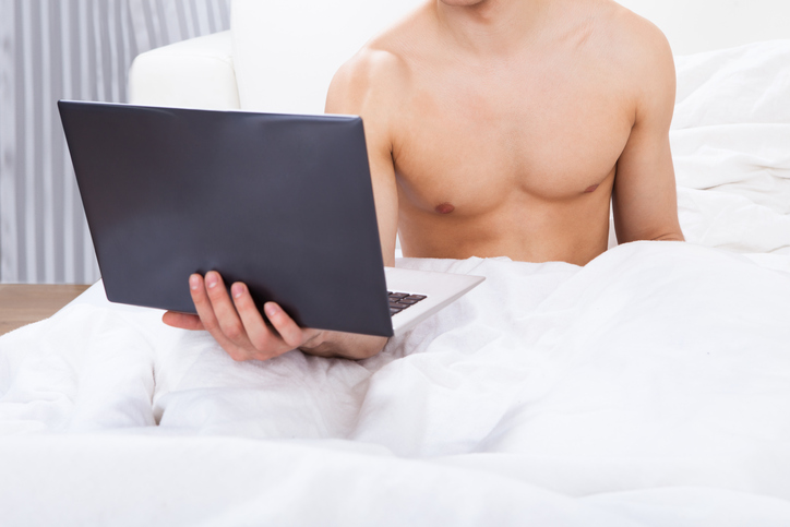 Midsection of shirtless man holding laptop in bed at home